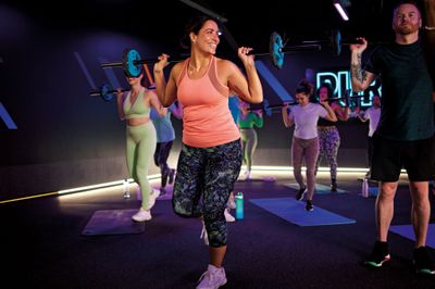 A smiling woman works out in a PureGym Pump class using weights. In the background other people are taking part in the class.